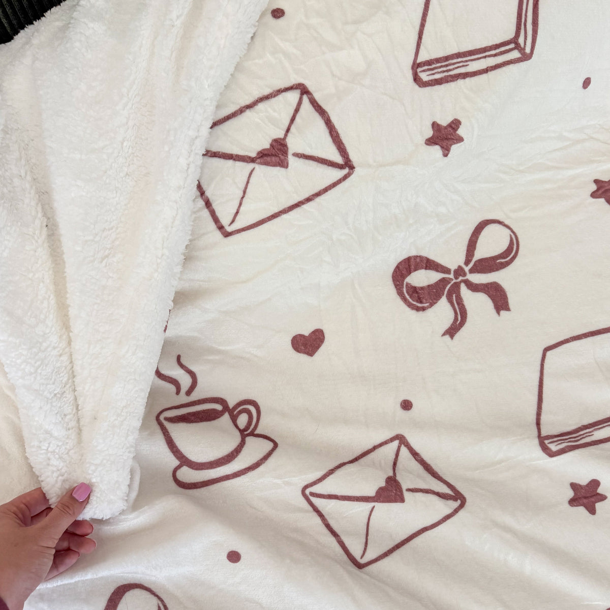 A close-up of a soft fleece blanket with a cream-colored sherpa backing and a printed pattern of dusty rose envelopes, bows, hearts, stars, and a coffee cup. A hand is visible holding the corner of the blanket to show its plush texture.