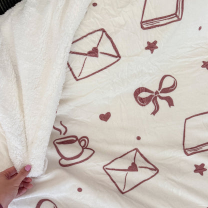 A close-up of a soft fleece blanket with a cream-colored sherpa backing and a printed pattern of dusty rose envelopes, bows, hearts, stars, and a coffee cup. A hand is visible holding the corner of the blanket to show its plush texture.
