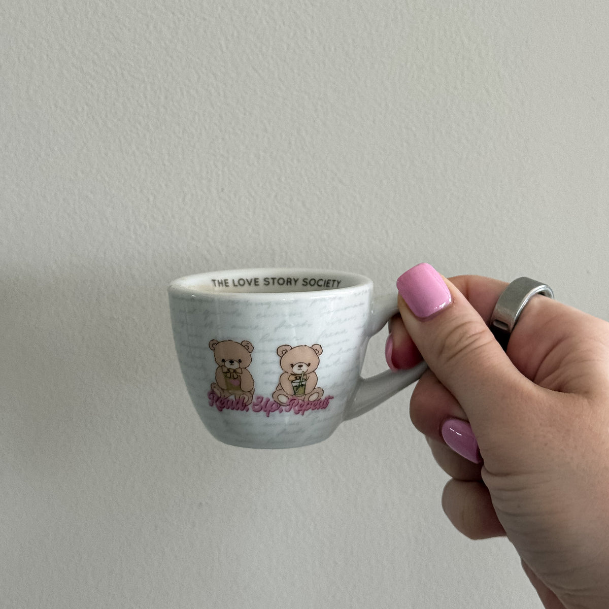 A hand with pink nail polish holds a small white ceramic coffee cup featuring two cartoon teddy bears and the text 'read, sip, repeat' against a plain off-white wall.