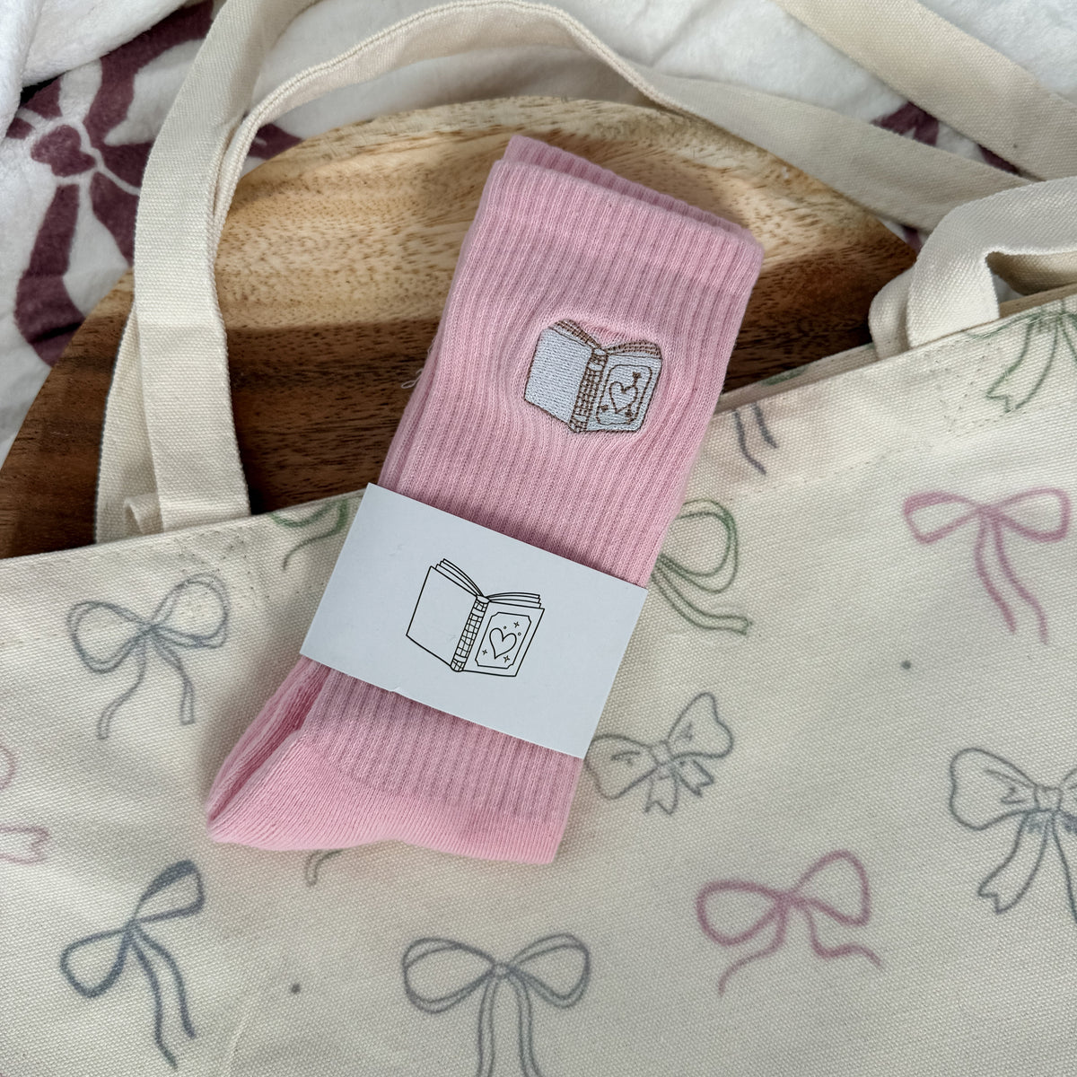 A pair of light pink ribbed socks with a small book graphic, wrapped in a white paper band, displayed on a cream canvas tote bag with bow patterns.