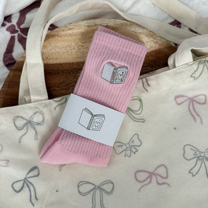 A pair of light pink ribbed socks with a small book graphic, wrapped in a white paper band, displayed on a cream canvas tote bag with bow patterns.