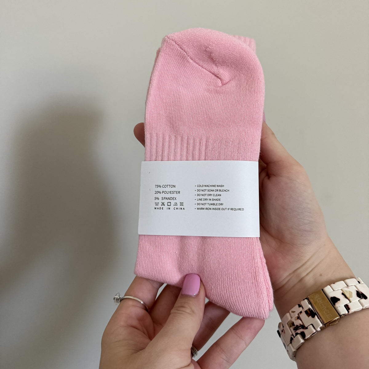 A pair of light pink crew socks held by two hands against a plain light gray wall. The socks have a white paper band wrapped around the middle with black text, and the person is wearing a patterned watch and a silver ring.